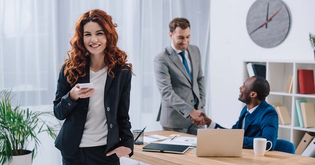 Eine junge Frau steht in einem Büro und ist glücklich über einen neuen Job, im Hintergrund schütteln sich zwei Männer die Hand | Immobilienverkauf bei Jobwechsel