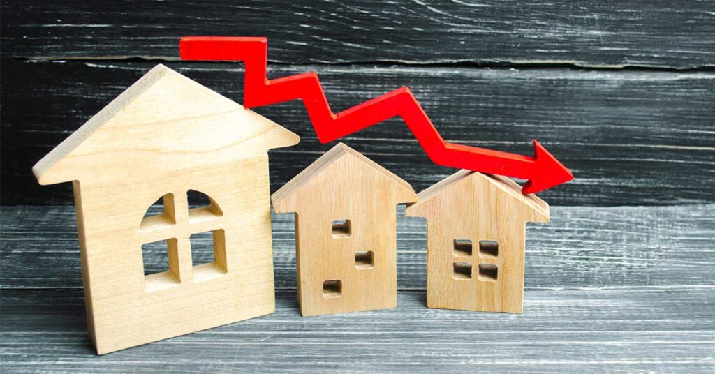 Drei unterschiedlich große Holzhäuschen stehen auf einem Holztisch vor einer Holzwand, über ihnen verläuft ein roter Pfeil im Zickzack nach unten | Preisstrategie Immobilienverkauf